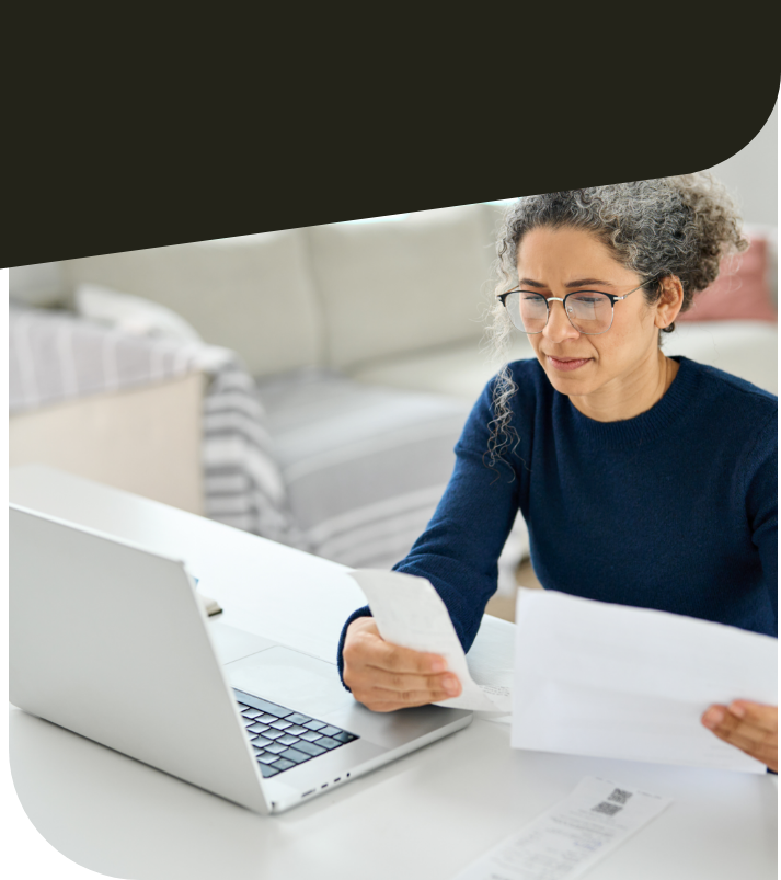 Mittelalte Frau sitzt an Schreibtisch. Sie blickt auf Zettel in ihrer Hand. Vor ihr steht ein Laptop.